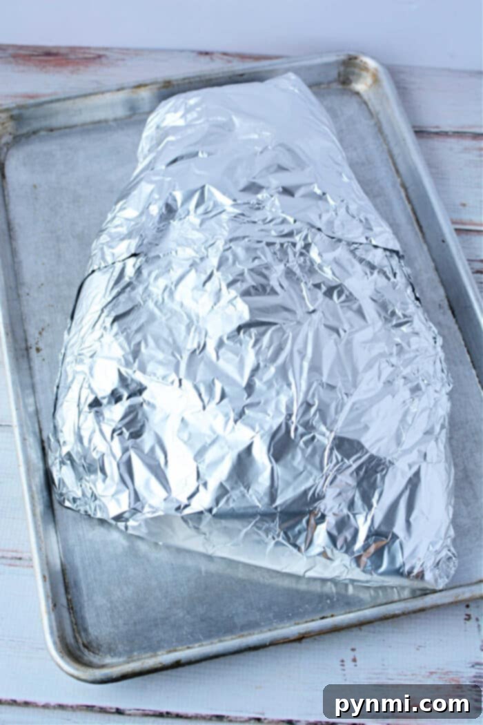 Perfect Fork-Tender Oven Brisket 6 A foil-wrapped brisket in the oven, rests on a baking sheet placed on a white wooden surface.