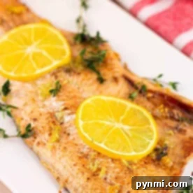 baked trout fillet garnished with lemon slices and thyme sits elegantly on a white plate, reminiscent of a dish youd find alongside baked trout. Nearby, a red-striped napkin adds a touch of charm.