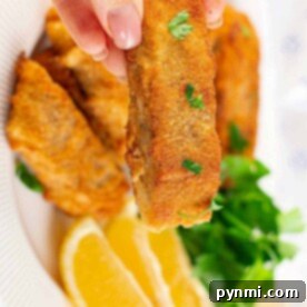 Golden Cornmeal Fish Fry 8 A hand holds a crispy fried piece of fish, perfectly coated in cornmeal for that extra crunch, over a plate adorned with lemon wedges and fresh greens.