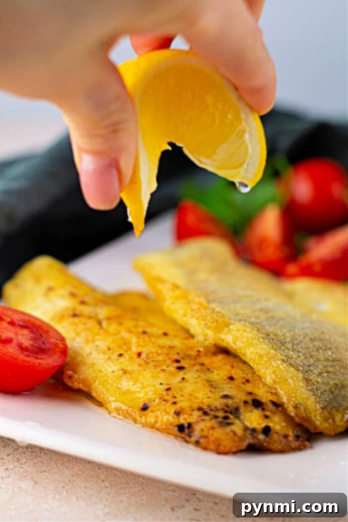 Hand squeezing lemon over seasoned fried Swai fish with cherry tomatoes on a white plate.