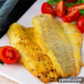 Two fried swai fish fillets rest on a white plate, adorned with sliced cherry tomatoes and a parsley garnish.