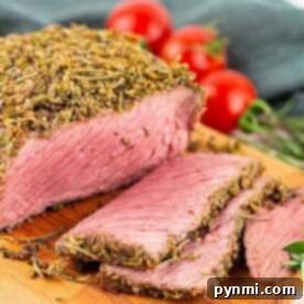 Sliced Bottom Round Roast with an herb crust rests on a wooden board, beautifully garnished with herbs and cherry tomatoes in the background.