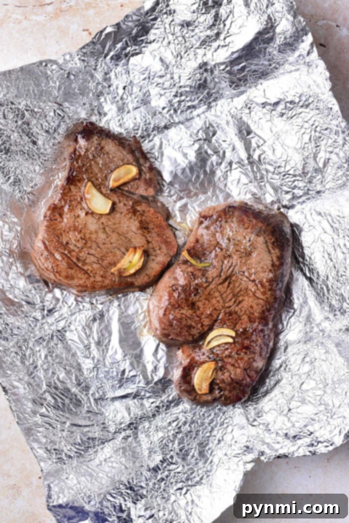 Two succulent cooked beef tenderloin medallions, rest on foil adorned with delicate garlic slices.