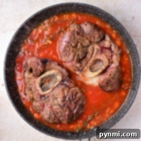 Beef shank with bone simmers in a pan with rich tomato sauce on a light granite surface.