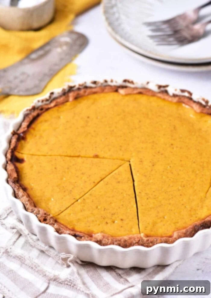 Velvet Southern Sweet Potato Dream 2 A golden-brown sweet potato pie in a white pie dish with a slice removed, revealing its smooth, orange filling. The pie rests on a patterned cloth napkin, with a silver serving utensil and plates in the background, ready to be enjoyed.