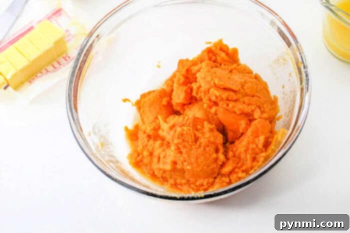Velvet Southern Sweet Potato Dream 4 A clear glass bowl brimming with perfectly mashed sweet potatoes, ready for baking. Beside it, a package of partially used butter and a small jar of vanilla extract are visible on a clean white surface, showcasing essential ingredients for a delicious sweet potato pie.