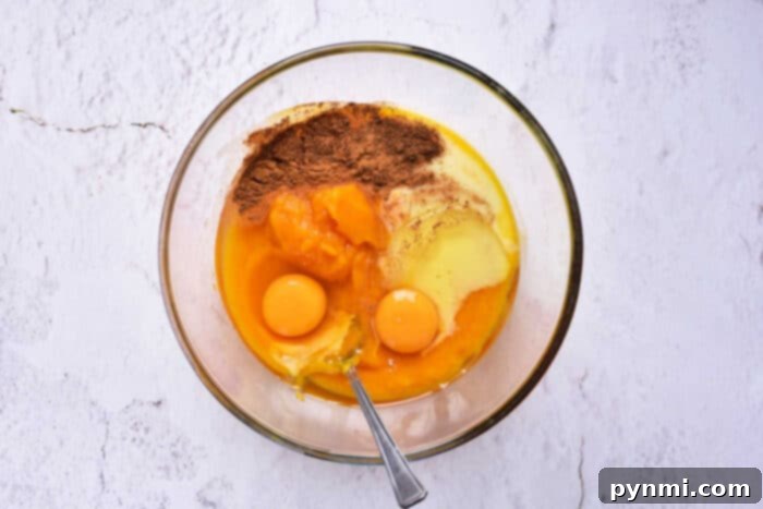 Velvet Southern Sweet Potato Dream 5 A large glass bowl on a white marble surface holds a creamy mixture of mashed sweet potatoes, sweetened condensed milk, and spices like cinnamon and nutmeg, with two raw eggs ready to be incorporated. A spoon rests inside the bowl.