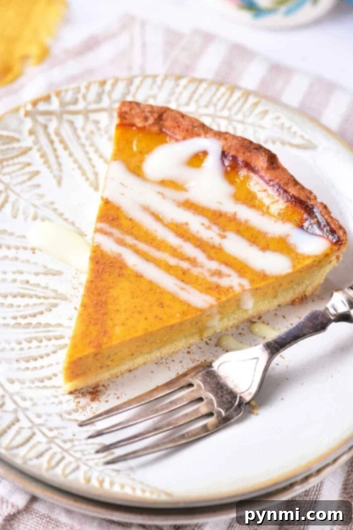 Velvet Southern Sweet Potato Dream 7 A beautifully sliced pumpkin pie topped with a delicate drizzle of white icing, presented on a decorative white plate. A silver fork rests beside the slice, inviting indulgence. The plate is positioned on a woven cloth, evoking the comforting essence of a classic sweet potato pie with sweetened condensed milk.