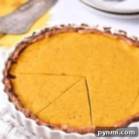 A sweet potato pie with a golden-brown crust and smooth filling sits in a white fluted pie dish. A slice has been cut and slightly separated from the rest of the pie, ready to be served. Plates, forks, and a pie server are visible in the background, set on a beige cloth.