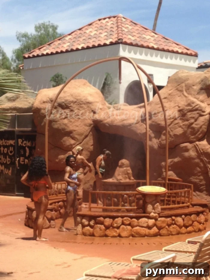 People in swimsuits stand around a mud bath area with rock formations and a wooden structure. In the background, there is a building with a terracotta-tiled roof. A sign reading "Welcome to Club Mud" is visible. A few lounge chairs are in the foreground, showcasing one of the best spas in Southern California.