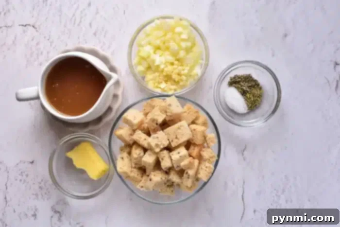Arrangement of fresh stuffing ingredients on a white textured surface, including a pitcher of golden broth, finely diced onions and garlic, a small bowl of mixed dried herbs and salt, a generous bowl of dried bread cubes, and a pat of butter on a plate.