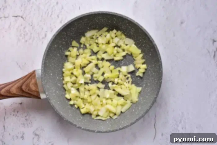 A close-up view of finely chopped onions sautéing in a non-stick frying pan with a wooden handle on a light kitchen counter, preparing aromatics for a savory stuffing recipe.