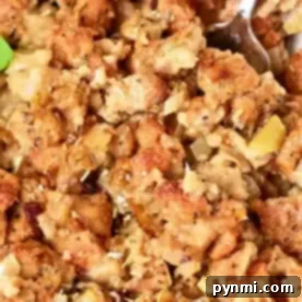 Close-up of a dish of oven-cooked stuffing, showcasing browned bread cubes mixed with cooked onions and herbs. The texture appears moist and chunky, with a spoon partially visible in the top right corner. Garnished with a small parsley leaf.