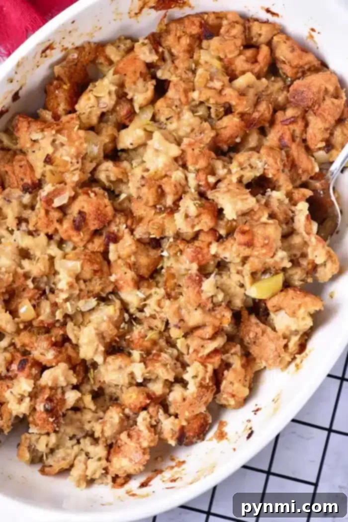A beautifully baked stuffing in a casserole dish, captured up close to highlight its golden-brown, crispy top and moist, flavorful interior, made with bread, herbs, and sautéed vegetables. A silver serving spoon is positioned to the right, ready for scooping.