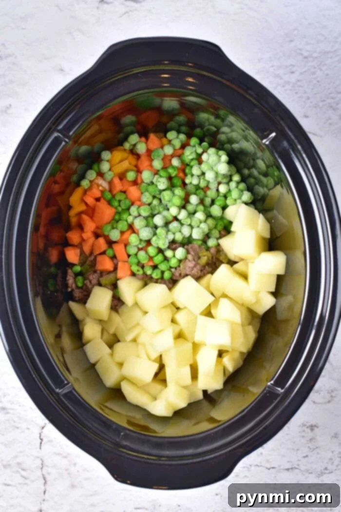 A crockpot brimming with diced potatoes, carrots, peas, and sliced onions sits on a light gray surface, ready to transform into a creamy potato and hamburger soup.
