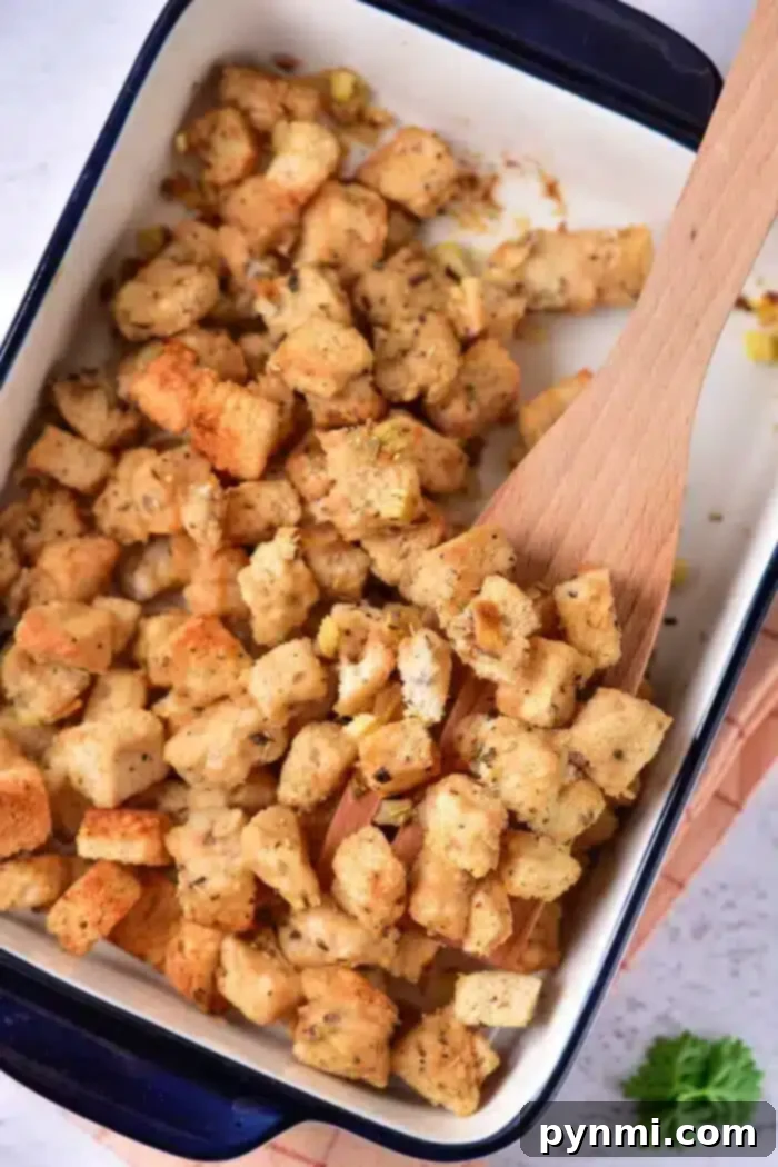 Golden Sourdough Croutons 2 A baking dish filled with evenly browned sourdough croutons, seasoned with herbs and spices. A wooden spoon rests among the croutons, while a sprig of parsley peeks from the bottom right corner outside the dish.