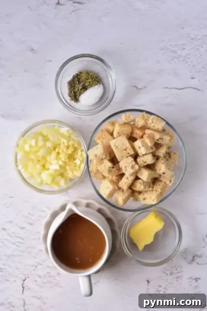 Golden Sourdough Croutons 3 Top-down view of ingredients for a stuffing recipe, including cubed sourdough croutons, chopped onions, minced garlic, a small dish of butter, a bowl of seasoning with salt and pepper, and a gravy boat of brown liquid on a light textured surface.