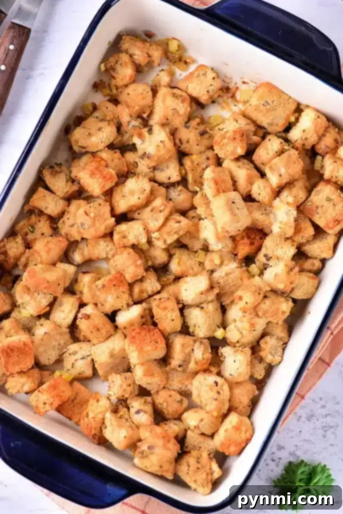 Golden Sourdough Croutons 7 A baking dish filled with seasoned sourdough croutons, possibly stuffing, arranged evenly. The dish is placed on a light-colored surface with a glimpse of a knife and a green herb nearby.