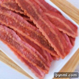 Slices of turkey bacon in the oven are arranged on a white tray atop a wooden board.