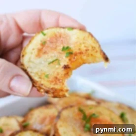 A close-up captures a hand holding a partially eaten crispy potato slice, topped with herbs, above a bowl of oven-roasted sliced potatoes.