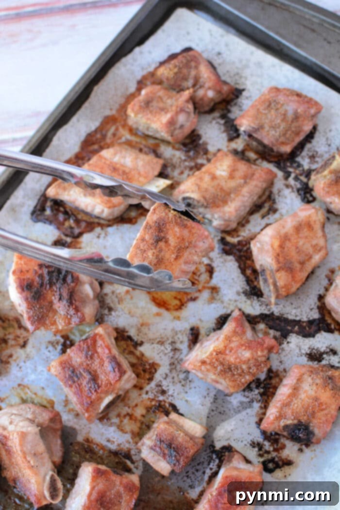 Tender Oven BBQ Riblets 6 Tongs holding a glistening, seasoned, and perfectly baked pork riblet, with more tender baked riblets in the oven visible on the tray.