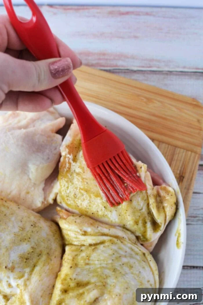 Crispy Skinned, Juicy Bone-In Chicken Thighs 7 A hand uses a red brush to spread marinade on raw chicken pieces in a white dish, prepping them before discovering how long to bake chicken thighs at 425 for perfect results.