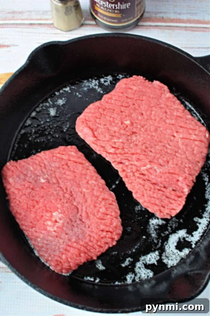 Two raw cube steak patties sizzling in a hot black cast iron skillet, demonstrating the initial stage of preparing tender cube steak.
