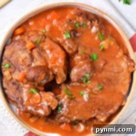 A plate of braised beef shin in rich tomato sauce with parsley garnish, resting on a napkin with handles in the background.