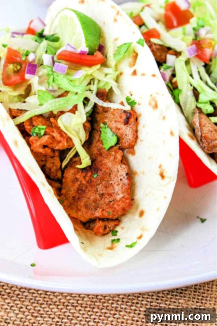 Mouthwatering Pork Tenderloin Tacos 2 A close-up of a Pork Tenderloin Taco filled with seasoned meat, shredded lettuce, chopped tomatoes, diced red onions, and cilantro, all inside a soft flour tortilla. The taco is placed in a red taco holder. A lime wedge is also visible on top of the filling.