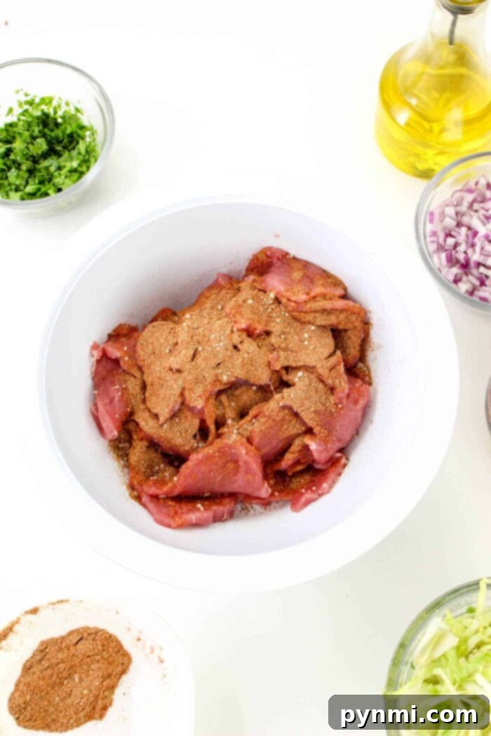 Mouthwatering Pork Tenderloin Tacos 5 A white bowl contains raw pork pieces seasoned with a brown spice mix, surrounded by bowls of chopped herbs, diced red onions, shredded lettuce, a small bowl of additional spices resembling the ones used in pork tenderloin tacos, and a bottle of olive oil on a white background.
