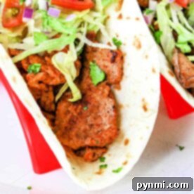 A close-up of a Pork Tenderloin Taco filled with seasoned meat, shredded lettuce, chopped tomatoes, diced red onions, and cilantro, all inside a soft flour tortilla. The taco is placed in a red taco holder. A lime wedge is also visible on top of the filling.