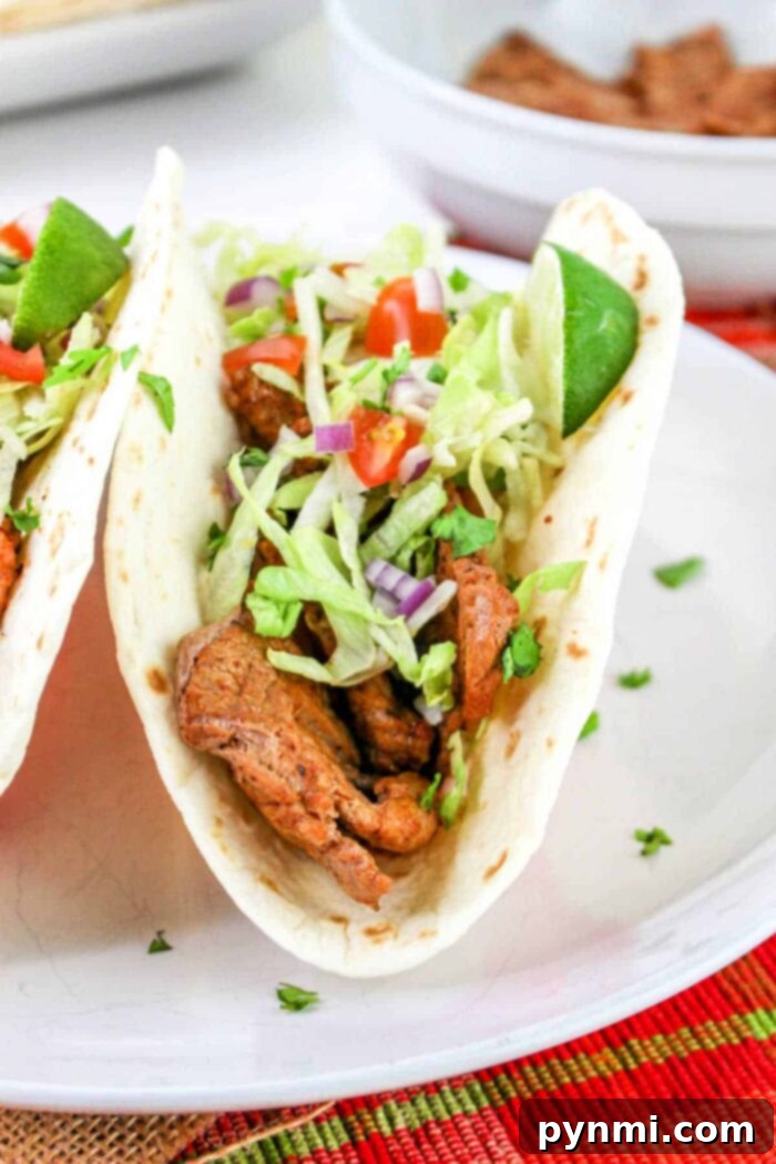Mouthwatering Pork Tenderloin Tacos 8 A close-up of a pork tenderloin taco filled with grilled pieces of meat, shredded lettuce, chopped tomatoes, and diced purple onions, garnished with a lime wedge. The taco is placed on a white plate with part of another taco visible in the background.