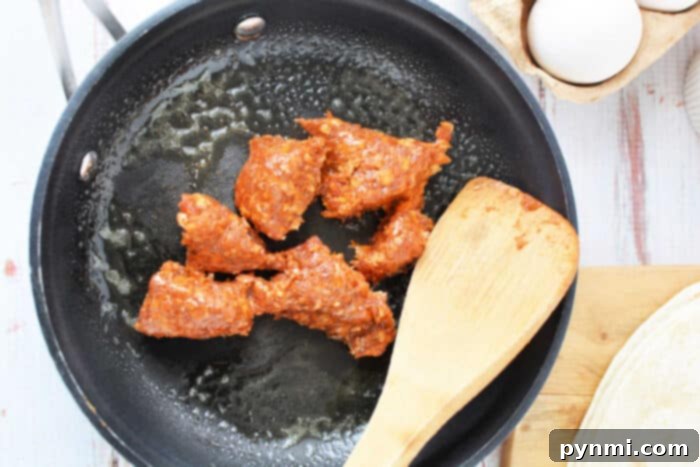 A black skillet sizzles with pieces of chorizo being cooked, a wooden spatula resting in the pan. Nearby on the counter, a carton of eggs awaits its turn, next to a tortilla on a wooden cutting board—perfect for learning how to cook chorizo and eggs.