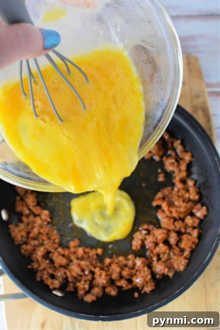 A hand is seen pouring whisked eggs from a bowl into a frying pan that contains browned ground meat, possibly chorizo. The pan rests on a wooden surface. The person is holding a whisk in the bowl, which is slightly tilted. This demonstrates how to cook chorizo and eggs effortlessly.