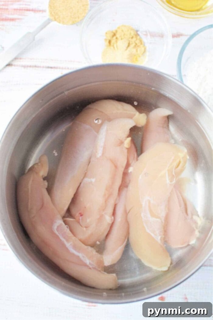 Raw chicken tenders seasoned with various spices, arranged in a silver bowl on a white textured background, highlighting the fresh ingredients before cooking and prompting thoughts on how to boil chicken tenders for optimal flavor and juiciness.
