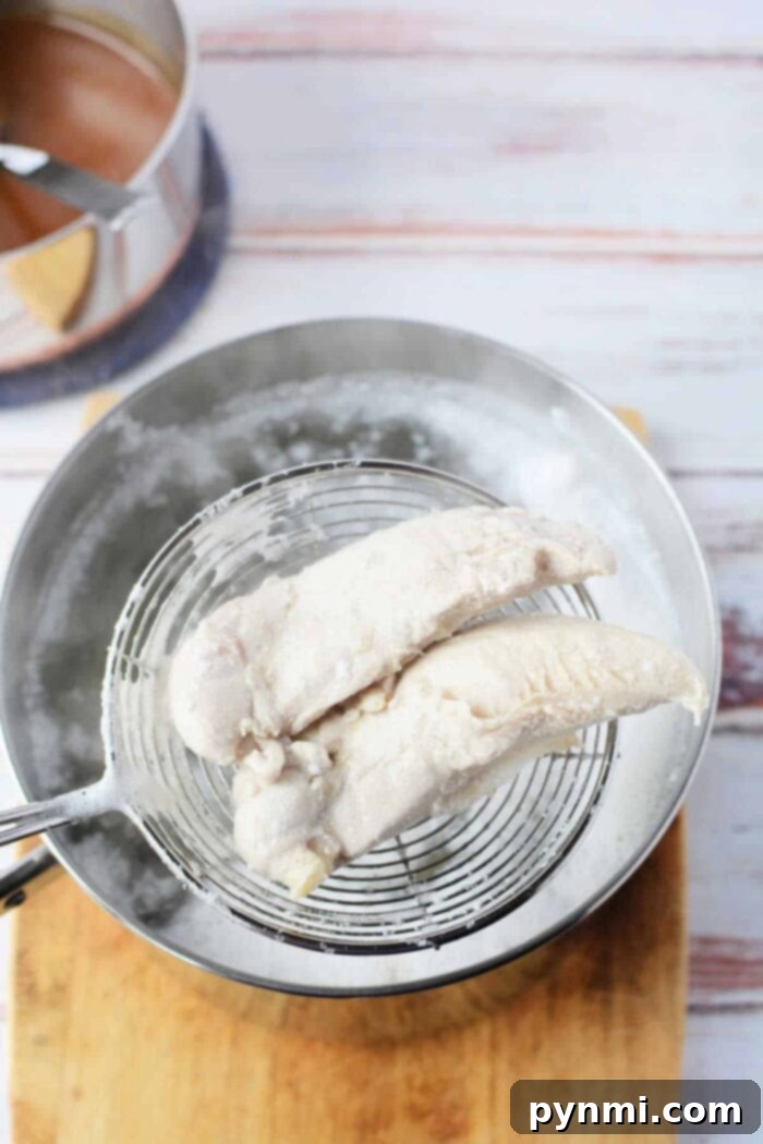 Two perfectly cooked chicken tenders being gently lifted from a steaming pot of water with a slotted spoon and placed onto a rustic wooden cutting board, illustrating the tenderness achieved by careful boiling.