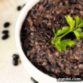 A delightful bowl of refried black beans recipe, topped with a fresh sprig of parsley, sits amid scattered black beans on the surface, inviting you to savor every bite.