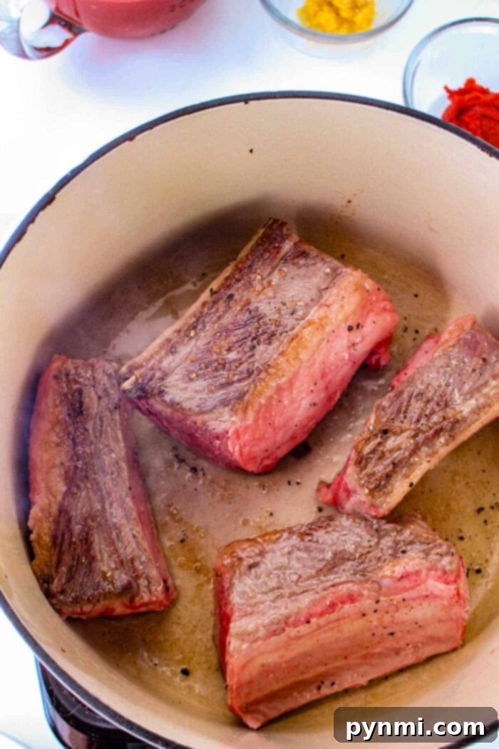 Four pieces of browned short rib meat are cooking in a large pot, perfect for tacos. The meat has a seared exterior. In the background, there are small bowls with ingredients, including a bright red and yellow substance. The pot and ingredients are set on a white surface.