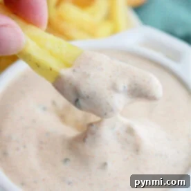 A hand dipping a fry into creamy, seasoned Blackened Ranch Sauce from Popeyes in a white bowl, with more fries in the background.