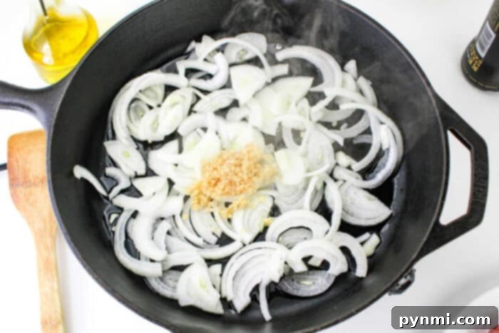 Unlocking Bratwurst Perfection 4 A robust cast iron skillet sits on a stovetop, filled with an aromatic mix of thinly sliced onions and fragrant minced garlic, with steam gently rising from the pan. A wooden spatula rests beside the skillet, ready for use, while a bottle of olive oil is visible in the soft background. This scene perfectly sets the stage for expertly preparing bratwurst, promising rich flavors and a delightful culinary experience.