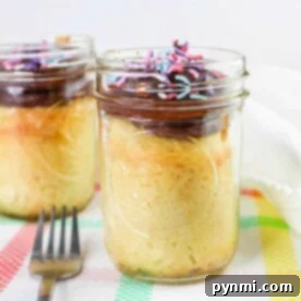 Vanilla cake in glass jars, showcasing an elegant take on how to bake cake in a jar, are adorned with chocolate frosting and colorful sprinkles while resting on a patterned tablecloth.