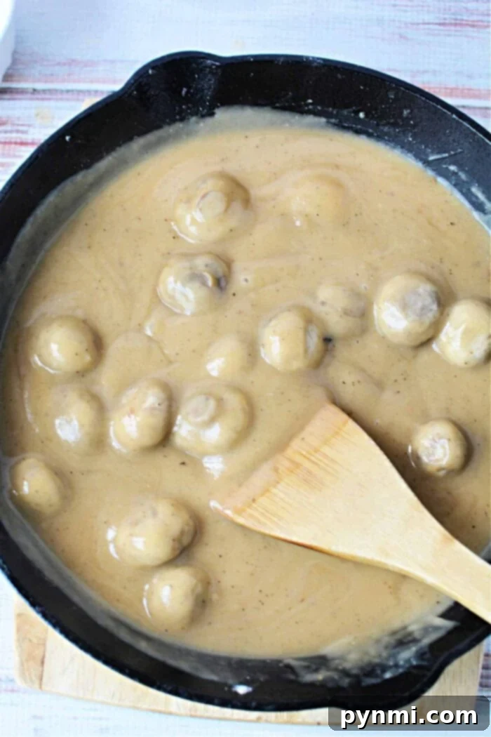 Velvety Mushroom Gravy 2 A skillet filled with succulent mushrooms smothered in a creamy brown sauce enriched with the flavors of cream of mushroom gravy, skillfully stirred with a wooden spatula.