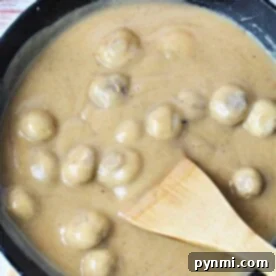 A skillet filled with succulent mushrooms smothered in a creamy brown sauce enriched with the flavors of cream of mushroom gravy, skillfully stirred with a wooden spatula.