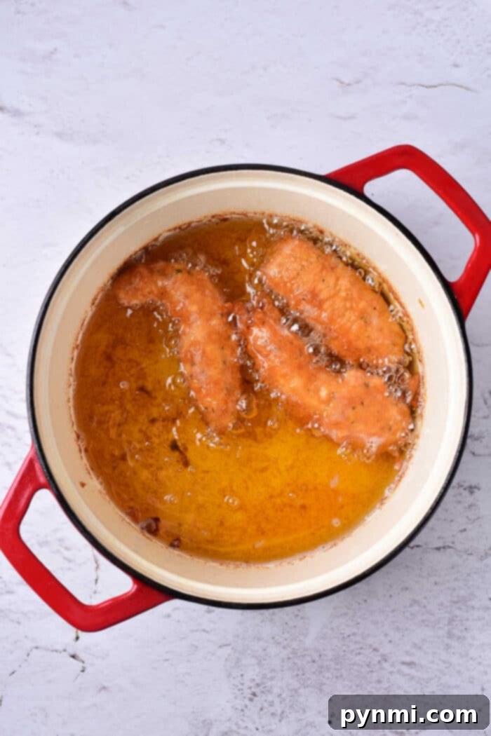 Two beer battered chicken tenders frying in hot oil alongside in a red-handled pot on a light gray surface.