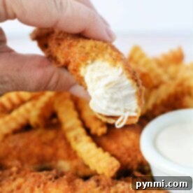 Savor the moment with a hand holding a crispy chicken tender, perfectly paired with crinkle-cut fries and a small cup of dipping sauce. Discover how to make chicken tenders that are just as golden and delicious right in your own kitchen.