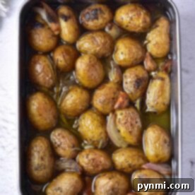A baking tray of confit roasted baby potatoes with herbs and garlic cloves rests on a white surface.
