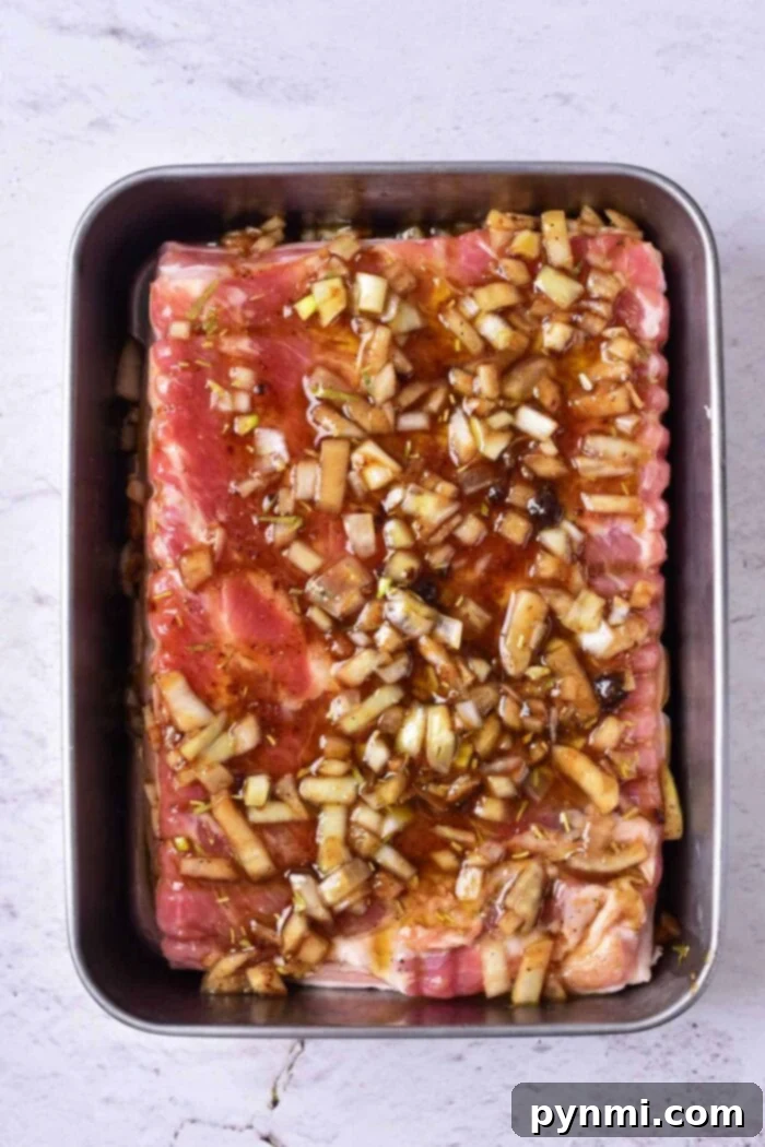 Raw pork belly, beautifully scored and generously coated with a rich marinade of chopped onions, garlic, and spices, resting enticingly in a rectangular metal tray on a light gray surface, perfectly prepped for a flavorful confit cooking experience.