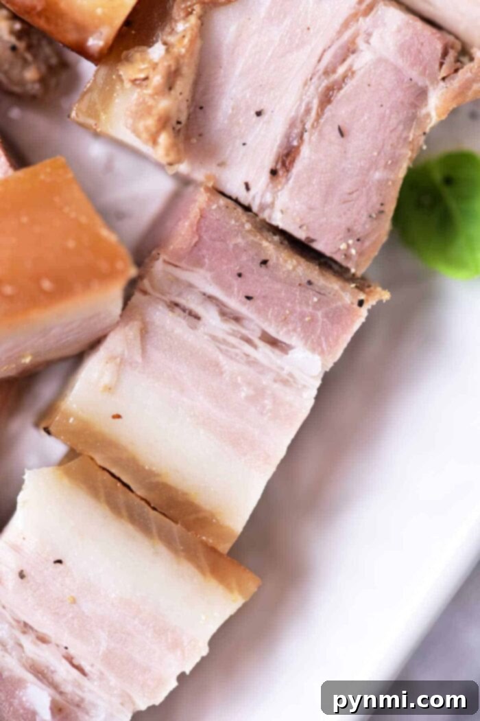 Close-up of perfectly cooked and sliced pork belly on a white plate, revealing beautiful layers of succulent meat and rich, rendered fat. This exquisite dish is the result of mastering the confit pork technique, garnished with fresh rosemary.
