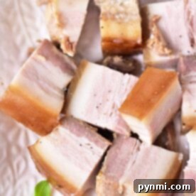 Learn how to confit pork and enjoy cubed pieces of layered cooked meat with luscious fat on a white dish, elegantly garnished with a small green sprig.