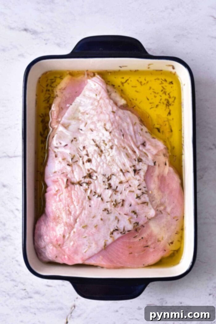 Mastering Turkey Breast Confit 4 A raw turkey breast marinating in oil and seasonings, destined for a delicious turkey confit, sits in a rectangular white and black roasting pan on a light-colored surface.